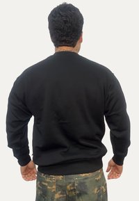 Uomo con capelli corti e ricci e tatuaggio sul collo indossa una felpa nera e pantaloni con motivo mimetico, vista di spalle con le braccia rilassate lungo i fianchi.