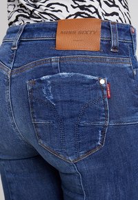 Vaqueros de mezclilla azul con un corte relajado, un parche de cuero "MISS SIXTY" y detalles desgastados en el bolsillo trasero.