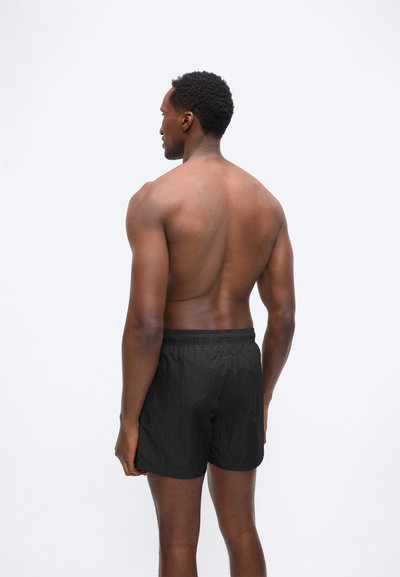 Άνδρας με κοντά σγουρά μαλλιά στέκεται γυμνή πλάτη, φοράει μαύρα υφασμάτινα σορτς, μπροστά σε απλό ανοιχτό φόντο.