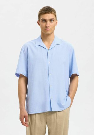 Camicia azzurra a maniche corte realizzata in tessuto testurizzato, con chiusura a bottone, colletto a punta e vestibilità rilassata. Abbinata a pantaloni beige.