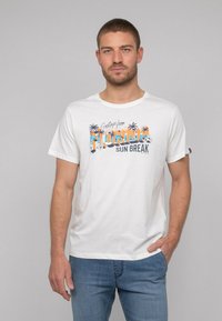 T-shirt en coton blanc avec l'impression "Salutations de Floride" en bleu et orange, présentant des graphiques de palmier. Manches courtes, coupe décontractée.