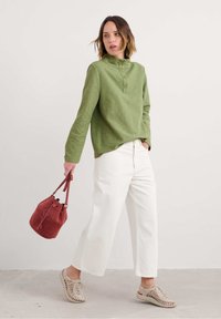 Sieviete, kas ģērbusies zaļā pogātā puloverī, baltās platās bikses, bēšās austās kedas, turot sarkanu samta iepirkumu somu, stāvot pretī vienkāršai sētiņai.