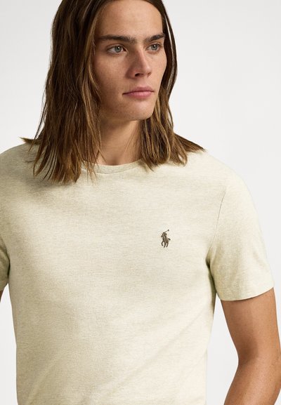 Světlé béžové tričko s krátkým rukávem z měkké texturované látky s malým hnědým vyšívaným logem na levé straně hrudi.