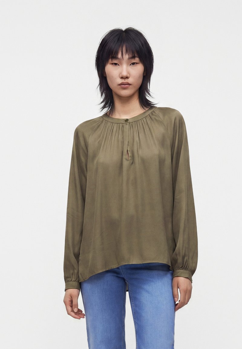 Jeune femme aux cheveux noirs portant une blouse ample à manches longues vert olive et un jean bleu, debout devant un fond clair uni.