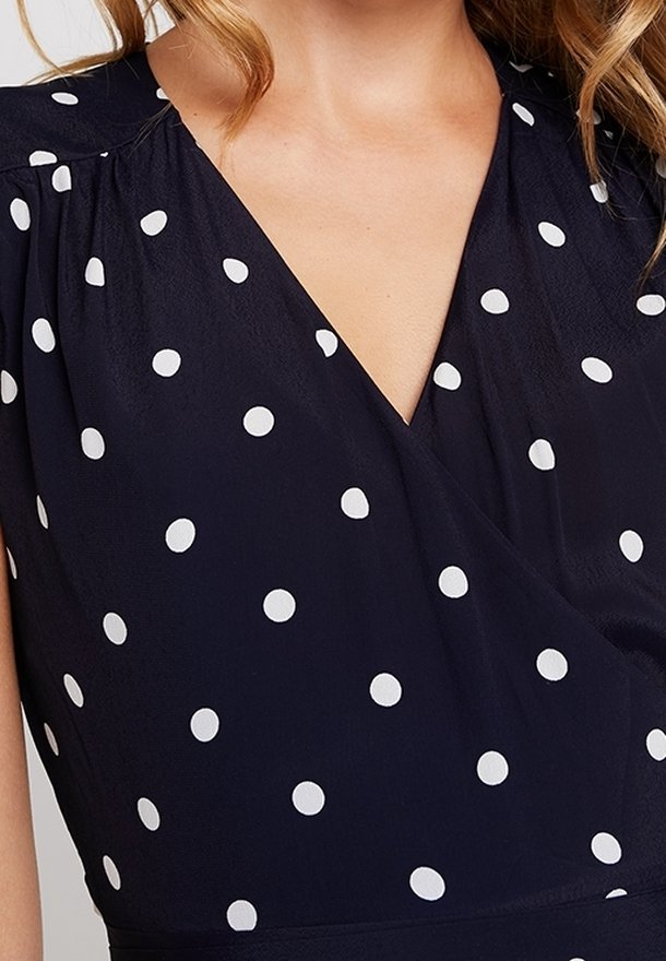 Granatowa sukienka w białe groszki, z dekoltem w serek i miękkim, lejącym się materiałem. Rękawy są marszczone na ramionach, co dodaje szczegółu.