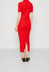 Robe en tricot rouge à manches courtes, texture côtelée, longueur mi-mollet, fente arrière, associée à des chaussures noires à talons carrés.