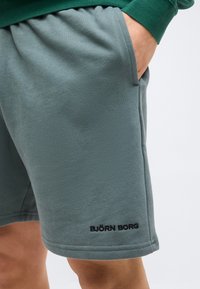 Άτομο που φοράει πράσινα σορτς με ελαστική μέση και πλαϊνή τσέπη, το χέρι εν μέρει μέσα στην τσέπη, μαύρο κείμενο λογότυπου κοντά στο τελείωμα.