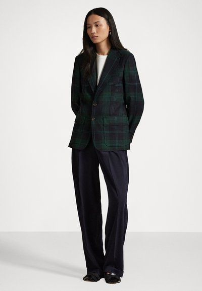 Žena stojaca v tmavozelenom a čiernom kockovanom sakách, bielom topánku, voľných čiernych nohaviciach a čiernych topánkach s mašľami na bielom pozadí.