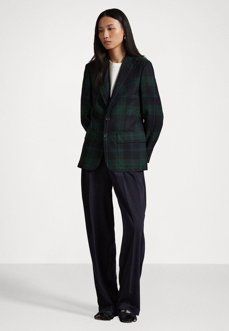 Donna in piedi che indossa un blazer a quadri verde scuro e nero, una maglietta bianca, pantaloni neri larghi e scarpe nere con fiocchi su uno sfondo bianco.