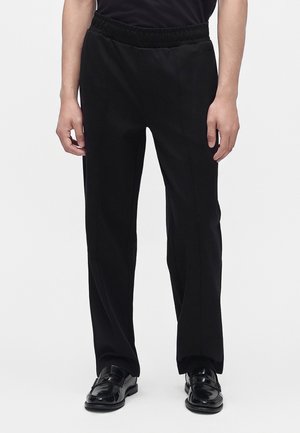Person wearing black elastic waist trousers and black polished dress shoes against a white background.