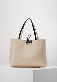 Sac cabas en cuir beige avec poignées noires, fermeture à cordon et petite plaque métallique avec le logo Guess au centre avant.
