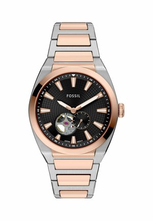 Montre Fossil avec un cadran rond noir, des accents en or rose et un bracelet en acier inoxydable argenté. Présente un mouvement squelettique et un motif texturé.