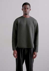 Pull noir à texture avec un motif en zigzag, col rond, et manches longues, porté avec un pantalon noir ample. Fond uni.