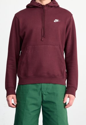 Mężczyzna w bordowej bluzie Nike z kapturem i przednią kieszenią oraz zielonych spodniach, stojący na białym tle.