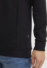 Sudadera negra con un bolsillo lateral, dobladillo y puños acanalados, textura suave y una pequeña etiqueta de la marca en la costura inferior. Combinada con unos vaqueros azules.