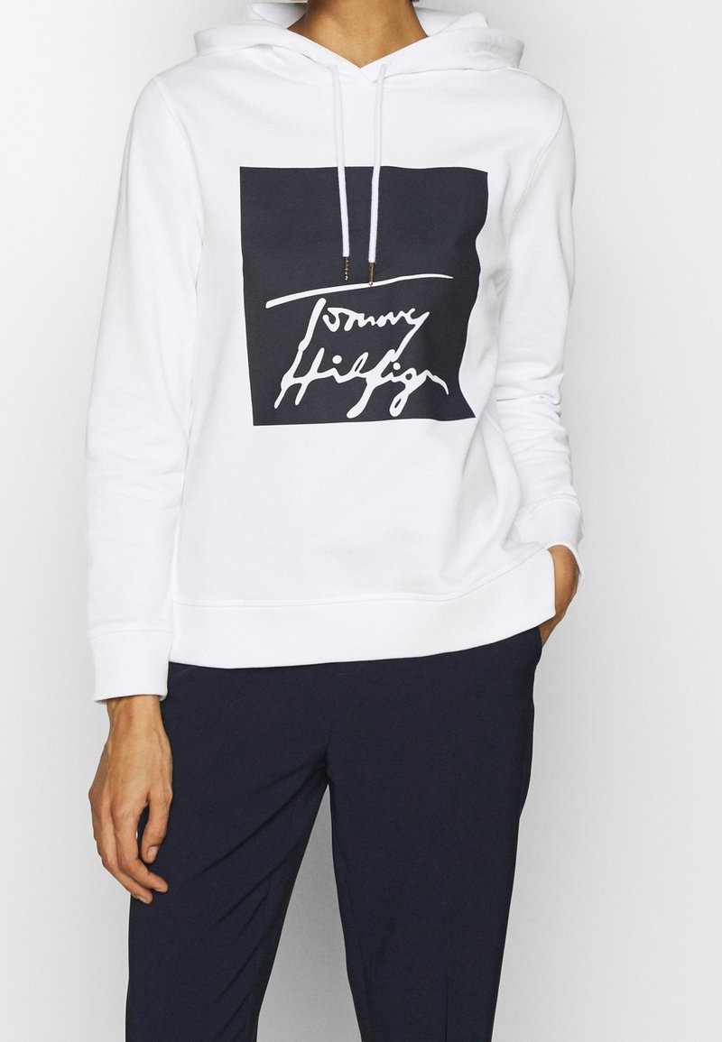 Sudadera con capucha blanca que presenta un cuadrado en azul marino con un logotipo de firma en blanco. Hecha de una tela suave, tiene un corte relajado y puños acanalados.