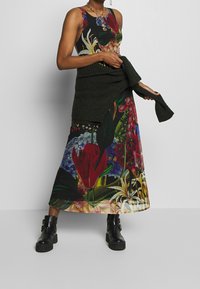 Kvinde iført en blomsterprint maxi-kjole med sorte ankelstøvler, som holder en mørkegrøn strikket sweater bundet om livet.