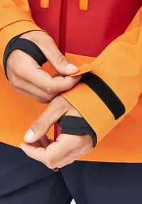 Pomarańczowa i czerwona kurtka ma kontrastowy czarny rzep u mankietu. Ręce regulują mankiet, pokazując gładką fakturę materiału i zarysowaną konstrukcję.