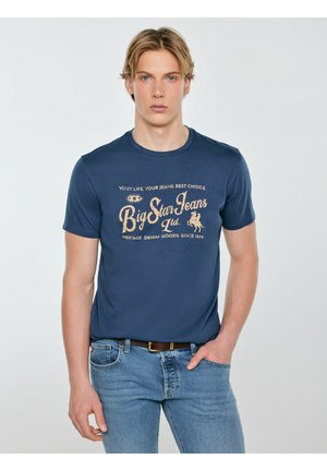 Młody mężczyzna o blond włosach, ubrany w granatowy t-shirt z napisem „Big Star Jeans Ltd.” oraz jasnoniebieskie jeansy, stojący z jedną ręką w kieszeni.