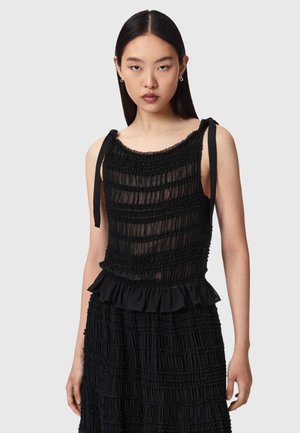 Femme aux cheveux raides et foncés porte un haut noir sans manches froncé avec des bretelles nouées et une jupe assortie sur un fond clair uni.