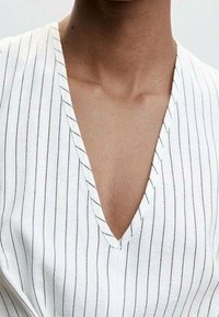 Persona vistiendo una camiseta blanca con cuello en V y finas rayas verticales negras, que muestra la zona del cuello y la parte superior del pecho.