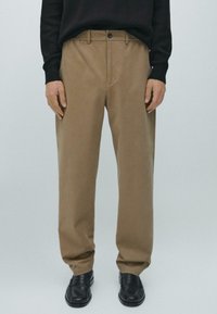 Hnědé chino kalhoty s rovnými nohavicemi, vyrobené z hladké tkaniny. Mají přední kapsy a zapínání na knoflík. Nošeny s černými botami.