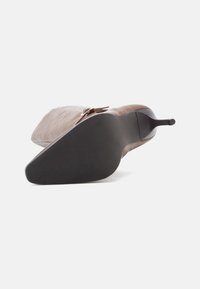 Καφέ ψηλοτάκουνη γόβα με λείο δέρμα στην επάνω μέρος, μυτερή μύτη και υφασμάτινη μαύρη λάστιχη σόλα. Διαθέτει διακοσμητική αγκράφα.