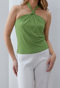 Žena v zeleném topu na ramínka a bílých kalhotách, stojící s rukama u těla proti jednobarevnému světlému pozadí.