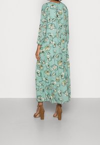 Mujer vistiendo un vestido largo floral en verde menta con mangas largas, de pie descalza frente a un fondo blanco liso.