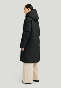 Svart lång kappa med huva, med en slät, matt yta och osynliga sömmar. Bärs över vida beige byxor och robusta svarta skor.