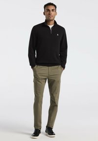 Schwarzer Reißverschluss-Pullover mit gerippter Textur, kombiniert mit olivgrünen Cargo-Hosen mit Seitentaschen und schwarzen Sneakers mit weißen Sohlen.