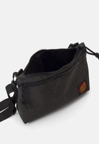 Black fabric crossbody bag with a zip closure, textured exterior, adjustable strap, and a red and yellow logo patch on the side.