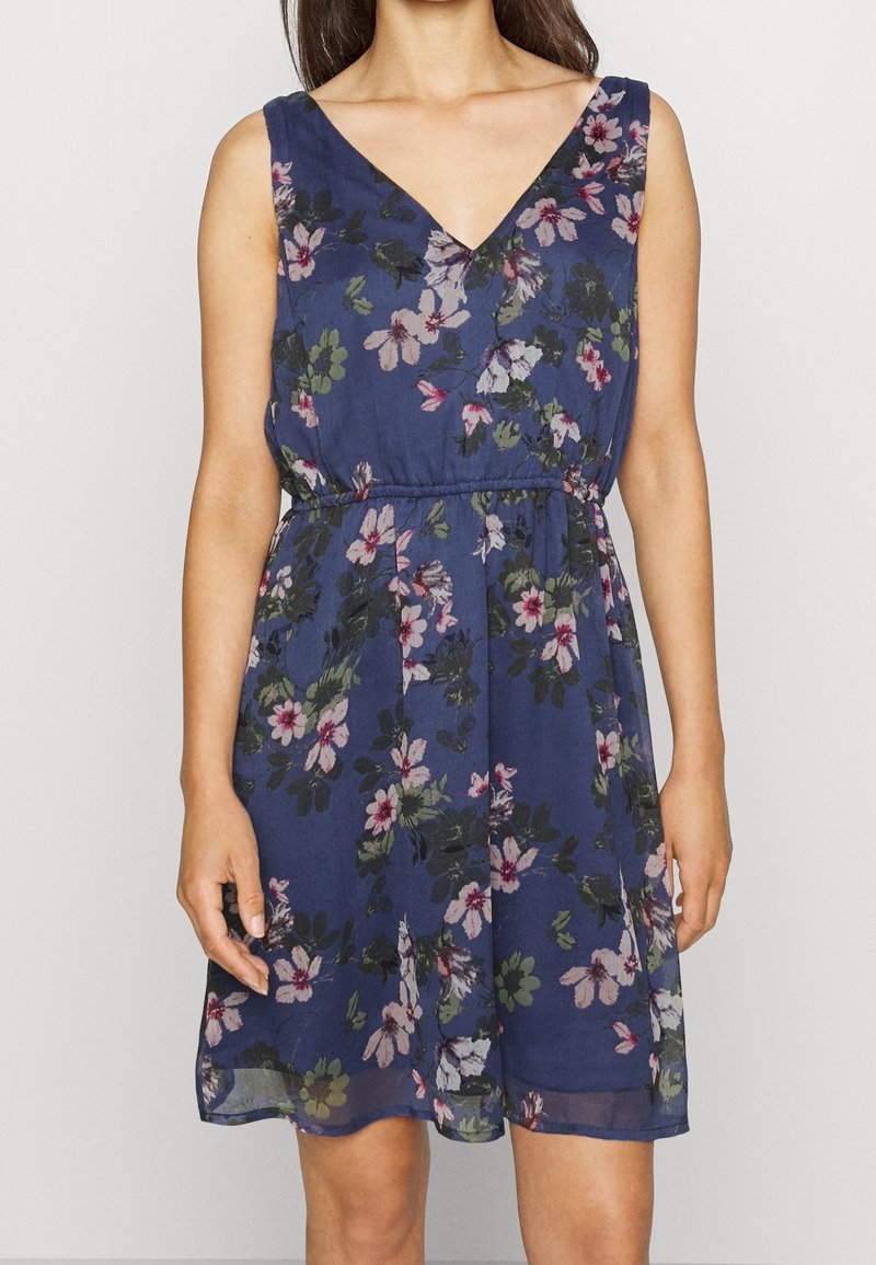 Femme portant une robe bleu marine sans manches avec des motifs floraux roses, blancs et verts et un décolleté en V.