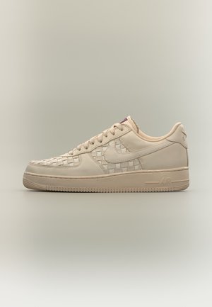 Baskets Nike Air Force 1 beige avec motif en cuir tissé sur les panneaux latéraux et semelle beige assortie sur fond neutre.