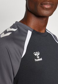 Nahaufnahme einer Person, die ein schwarz-graues Sportshirt mit weißen Streifen und dem weißen Hummel-Logo auf der Brust trägt.