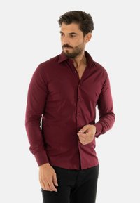 Camicetta a maniche lunghe di colore bordeaux con bottoni, caratterizzata da un colletto classico, un design aderente, una texture liscia e una vestibilità slim. Esibita con pantaloni neri.