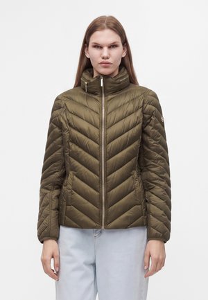 Femme aux cheveux longs et raides portant une veste matelassée vert olive avec un col montant et un pantalon bleu clair sur un fond uni.