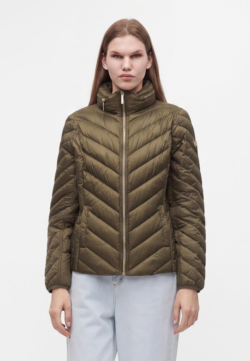Femme aux cheveux longs et raides portant une veste matelassée vert olive avec un col montant et un pantalon bleu clair sur un fond uni.