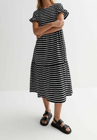 Mekko, jossa on musta-valkoisia raitoja, lyhyet röyhelösateet ja kerroksellinen hame, yhdistettynä mustiin tasapohjaisiin sandaaleihin. Yksinkertainen, rento muotoilu.