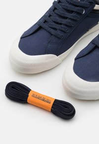 Baskets en toile bleu marine avec semelles en caoutchouc blanches, embouts texturés et lacets bleu foncé ; comprend un lacet bleu marine avec une étiquette orange.