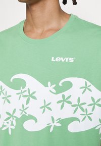 Homme portant un t-shirt vert Levi's avec des motifs blancs de vagues et de fleurs étoilées sur la poitrine.