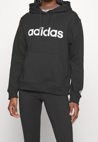 Czarna bluza z kapturem wykonana z mieszanki bawełny, z białym logo „adidas” na przodzie, kapturem ze sznurkiem oraz kieszenią kangur.