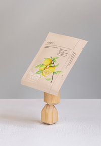 Väska med en beige bakgrund som har gula citroner, gröna blad och texten "Balans din skönhet", hålls upprätt av en trägeometrisk ställning.