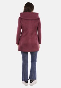 Cappotto bordeaux con colletto ampio e vestibilità sartoriale, realizzato in tessuto liscio. Abbinato a jeans a campana e sneakers bianche.
