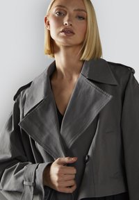 Lyhyt takki pehmeässä harmaassa kankaassa, jossa on isot kaulukset, yksi napitus ja sivutaskut. Rakenne on selkeä ja malli rento.