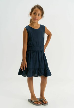 Pige med krøllet hår iført en ærmeløs marineblå kjole og grå flip-flops, stående med den ene hånd på hoften mod en hvid baggrund.