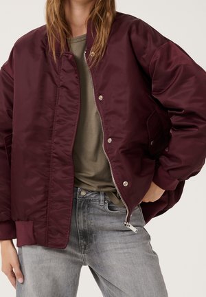 Femme portant une veste bomber bordeaux sur une chemise vert olive et un jean gris clair, debout avec une main dans la poche.
