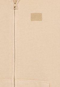 Bežna zip-up dukserica izrađena od teksturirane tkanine; sadrži mali, vezeni geometrijski logotip na lijevoj strani i zatvaranje na prednjem dijelu sa patentnim zatvaračem.