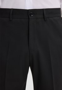 Schwarz maßgeschneiderte Hose mit Knopfverschluss, Gürtelschlaufen und einem weißen Hemd, das in die Taille gesteckt ist.