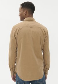 Rudas medvilnės ilgomis rankovėmis marškiniai, su sagomis ant rankogalių, nugaros papuošimu ir kreivu apvadu, dėvimi su mėlynomis džinsinėmis kelnėmis, ant paprasto balto fono.